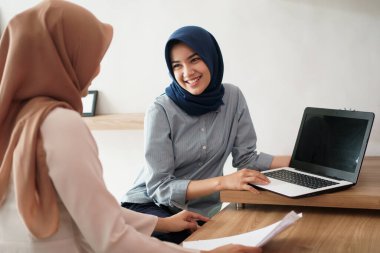 Çekici, genç Müslüman iş kadını laptopta çalışıyor ve masadaki modern ofisinde arkadaşıyla otururken gülümsüyor.