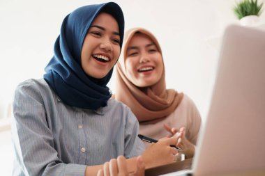 Çekici, genç Müslüman iş kadını laptopta çalışıyor ve masadaki modern ofisinde arkadaşıyla otururken gülümsüyor.