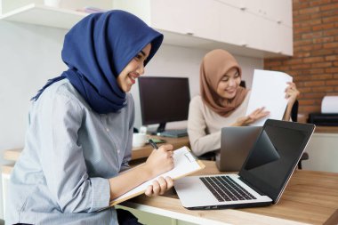 Çekici, genç Müslüman iş kadını laptopta çalışıyor ve masadaki modern ofisinde arkadaşıyla otururken gülümsüyor.