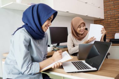 Çekici, genç Müslüman iş kadını laptopta çalışıyor ve masadaki modern ofisinde arkadaşıyla otururken gülümsüyor.