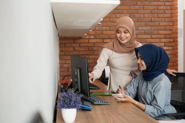 Attraente allegra giovane donna d'affari musulmana che lavora al computer e sorride mentre si siede alla sua scrivania ufficio moderno con la sua amica — Foto Stock