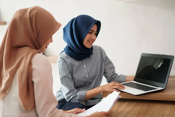 Attrayant joyeux jeune femme d'affaires musulmane travaillant sur ordinateur portable et souriant tout en étant assis à son bureau moderne avec son ami — Photo