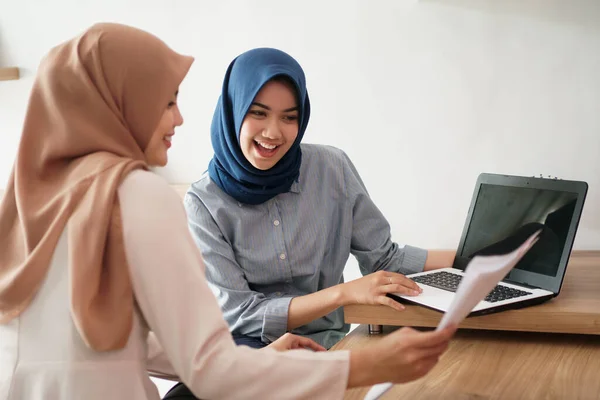 Atractiva joven mujer de negocios musulmana alegre trabajando en el ordenador portátil y sonriendo mientras se sienta en su escritorio oficina moderna con su amigo — Foto de Stock