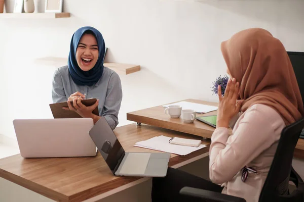 Attractive cheerful young muslim business woman working on laptop and smiling while sitting at her desk modern office with her friend — Stock Photo, Image