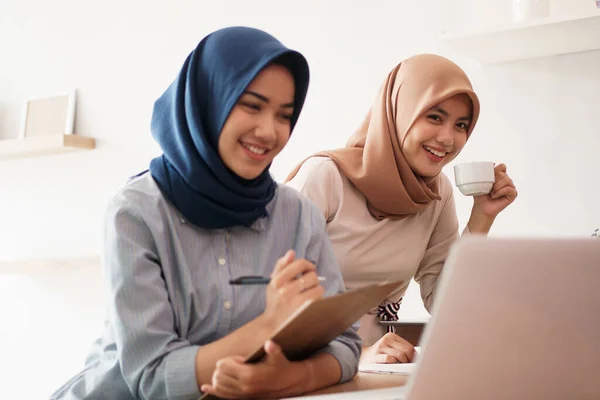 Atractiva joven mujer de negocios musulmana alegre trabajando en el ordenador portátil y sonriendo mientras se sienta en su escritorio oficina moderna con su amigo — Foto de Stock