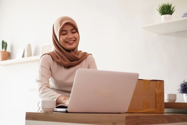 attractive cheerful young muslim business woman working on laptop and smiling while sitting at her desk modern office