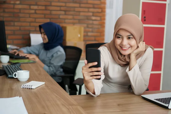 Atraente e alegre jovem muçulmano mulher de negócios está trabalhando em um computador e sorrindo enquanto sentado em uma moderna mesa de escritório com sua amiga que está tomando selfie — Fotografia de Stock