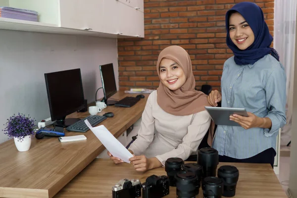 Attractive cheerful young muslim business woman smiling while sitting at her desk modern office with her friend — Stock Photo, Image