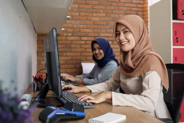 Приваблива весела молода мусульманська бізнес-леді, що працює на комп'ютері і посміхається, сидячи за її столом сучасний офіс зі своїм другом — стокове фото
