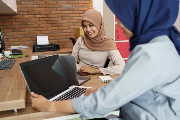 Atraente alegre jovem muçulmano mulher de negócios trabalhando no laptop e sorrindo enquanto sentado em sua mesa escritório moderno com seu amigo — Fotografia de Stock