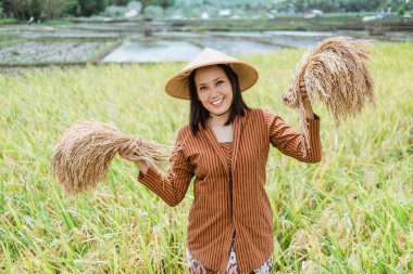 Asyalı kadın çiftçiler ayakta dikilmiş pirinç bitkileri taşıyan bir şapka takarken gülümsüyorlar.