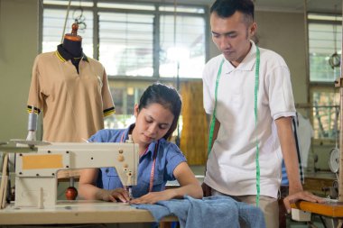 Erkek terzi yanında durur ve kadın terzinin dikiş makinesini kullandığını görür.