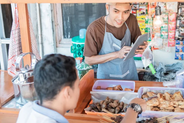 Angkringan jovem vendedor sorri usando tablet pc ao servir o cliente — Fotografia de Stock