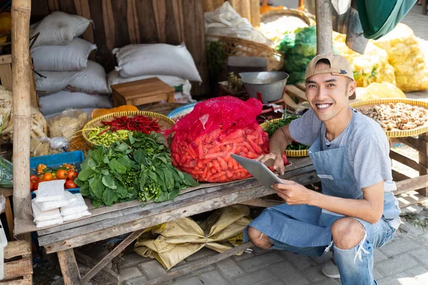 Ázsiai férfi árul zöldséges standot mosolyog, ahogy guggol egy tabletta segítségével — Stock Fotó