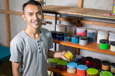 Erkek işçi, sarı boya şişesini rafın önünde tutarken gülümser.