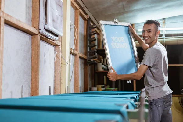 smiling young man holding the silkscreen frame and carefully observing the result of the film on the silkscreen