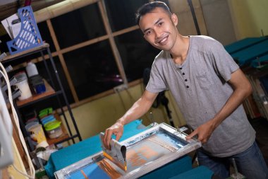 Genç Asyalı erkek işçi, tişört ekranı yazdırmak için ekran baskı çerçeveleri hazırlarken gülümsüyor