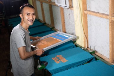 Erkek işçiler tişörtleri bastıktan sonra ekranın baskı çerçevelerini tutarken ve kaldırırken gülümsüyorlar