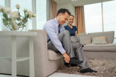 Bir erkek ayakkabılarını giyerken bir kadın şık bir oturma odasında eğleniyor..