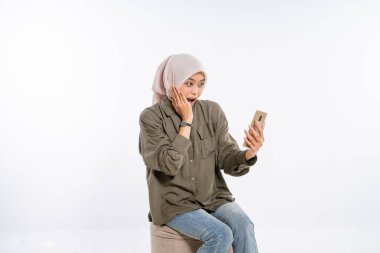 Başörtüsü takan şaşırmış bir kadın heyecanla ve merakla telefonuna bakıyor.