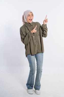 Zeytin gömleği ve kot pantolon giymiş neşeli genç bir kadın heyecanla işaret ediyor.