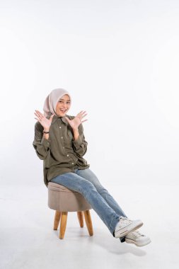 Şık tesettürlü neşeli bir kadın bir taburede oturur, gülümsemesiyle mutluluk saçar.