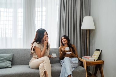 İki arkadaş gülüyor ve sıcak çayı paylaşıyor. Parlak, rahat bir yaşam alanında, neşe yaratıyor.