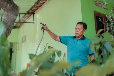 Bir adam aktif olarak bitkileri canlı, yemyeşil bir bahçede hortumla suluyor.