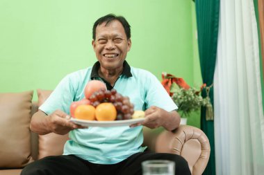 Neşeli yaşlı bir adam rahat bir ortamda canlı bir meyve tabağıyla mutlu bir şekilde oturur.