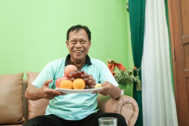 Yaşlı bir adam elma ve üzüm gibi renkli bir tabak meyve gösterirken gülümser.