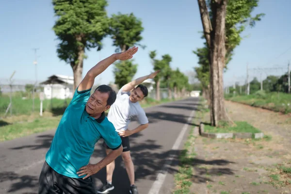 İki adam, güneşli bir günde, açık havada yan yana esneme hareketleri yaparken görülebilir.
