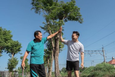 Güneşli bir parkta neşeyle beşlik çakan iki adam, dostluk ve zindeliği kutluyorlar.