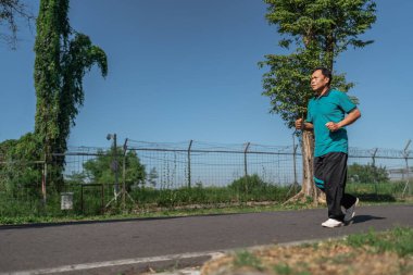 Bir adam yemyeşil ve açık mavi gökyüzüyle çevrili güzel bir yolda durmadan koşuyor.