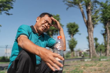 Dışarıda oturan yorgun bir adam, elinde bir su şişesi var ve gözlerini güneşten koruyor..