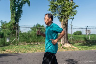 Yaşlı bir adam güneşli bir günde güzel ağaçlarla çevrili huzurlu bir parkta enerjik bir şekilde koşar.