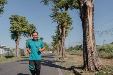 Neşeli ve hayat dolu bir adam ağaçlarla çevrili güneşli, güzel bir yolda enerjik bir şekilde koşar.