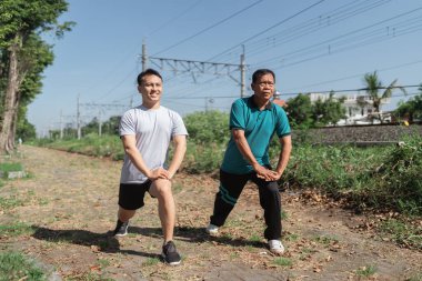 Tren raylarının oradaki manzaralı bir alanda iki adam hareketli gerinme egzersizleri yapıyor.