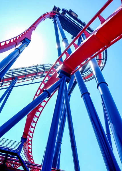 looking up to the high metal blue and red frame work of a high modern rollercoaster forming graphical shapes and lines, blue sky and blue pool, roller coaster construction is metal painted red and blue