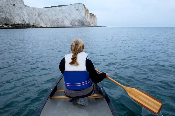 The back view of a sixty year old woman sitting in the front of a Kayak paddling with a wooden paddle she is wearing a life jacket and has blond hair with a pony tail going down the back of her life jacket around her is a flat calm blue sea and sky o