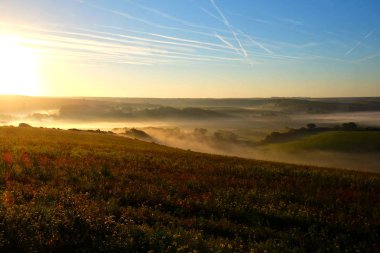 Cuckmere valley, East Sussex, United Kingdom, UK, covered in mist in the lower part of the valley with clear green rolling hills above with the sun shining on themthe sun is shining in the top left corner and there is an orange glow in the sky clipart