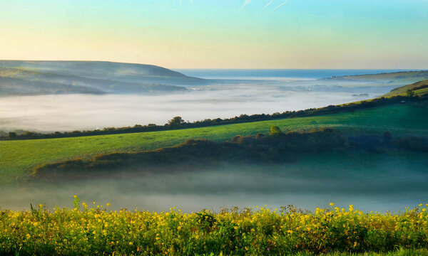 Cuckmere Valley, East Sussex, United Kingdom, UK, covered in mist in the lower part of the valley with clear green rolling hills above with the sun shining on them the sea is in the background with a orange glow in the blue sky
 