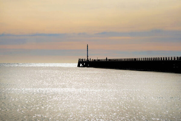 Newhaven, East Sussex, UK, United Kingdom, harbour wall jutting out from the right hand side, it is silhouetted black with the silhouettes of people fishing on the end the sea infront is glistening silver from the sun and the sun has set and the sky 