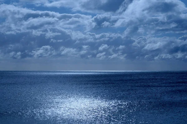 海和天空的下半部分是平静的深蓝色的大海 地平线上是一条白色的发光光芒 从月亮上半部分是天空中蓬松的云覆盖着月光下的蓝色光芒 — 图库照片