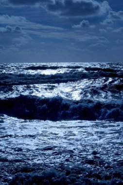 Şişme deniz dalgaları ile koyu mavi deniz karanlık gölgeler vardır burada dalgalar şişme ve güneş ışığı parlak beyaz ön planda ufukta denizin üzerinde olduğu bir karanlık Çakıl Plajı, dikey format üst yarısında gökyüzü kaplıdır