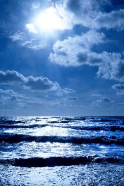 Şişme deniz dalgaları ile koyu mavi deniz burada dalgalar şişme ve güneş ışığı parlak beyaz üzerinde karanlık gölgeler vardır ön planda ufukta denizin karanlık pebble Beach, dikey çizgidir üst yarısında biçimidir gökyüzü cove
