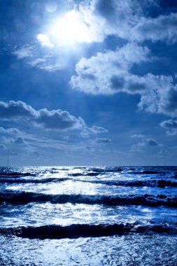 A dark blue sea with waves swelling the sea has dark shadows where the waves are swelling and the sunlight is glistening white on the sea on the horizon in the foreground is line of dark pebble beach, Vertical format in the upper half is the sky cove clipart