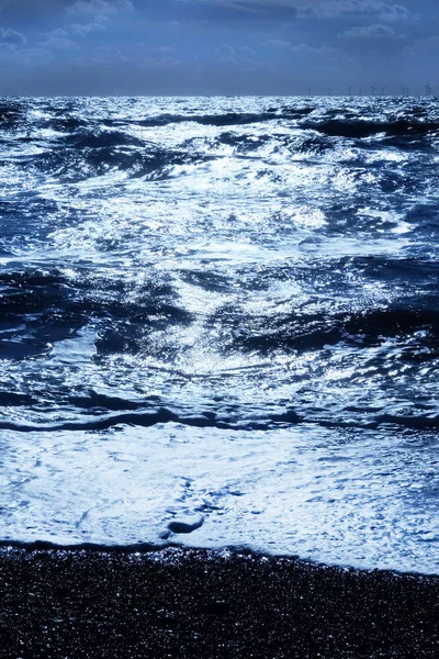 A dark blue sea with waves swelling the sea has dark shadows where the waves are swelling and the sunlight is glistening white on the sea on the horizon in the foreground is a dark pebble beach, Vertical format in the upper half is the sky covered in