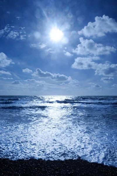 A dark blue sea with waves swelling the sea has dark shadows where the waves are swelling and the sunlight is glistening white on the sea on the horizon in the foreground is a dark pebble beach, Vertical format in the upper half is the sky covered in