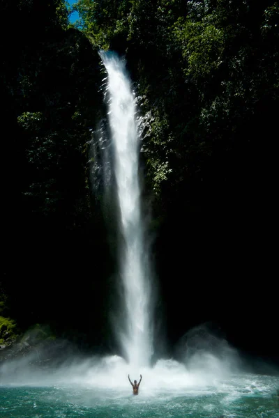 一个人站在瀑布底部的水池里 双手举在空中 显示出这个人和广阔的瀑布之间的规模差异 70米长的透明白水丝带 通过一个纯粹的 倾泻而过 — 图库照片