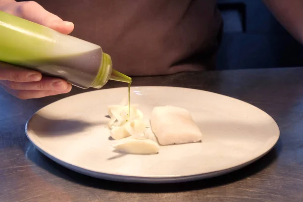 Close up of male chefs hands pouring green sauce from a clear plastic bottle onto a white plate with white fish on it, the plate is on a silver metal kitchen top behind is the part of the grey apron the chef is wearing in a fine dining restaurant.
